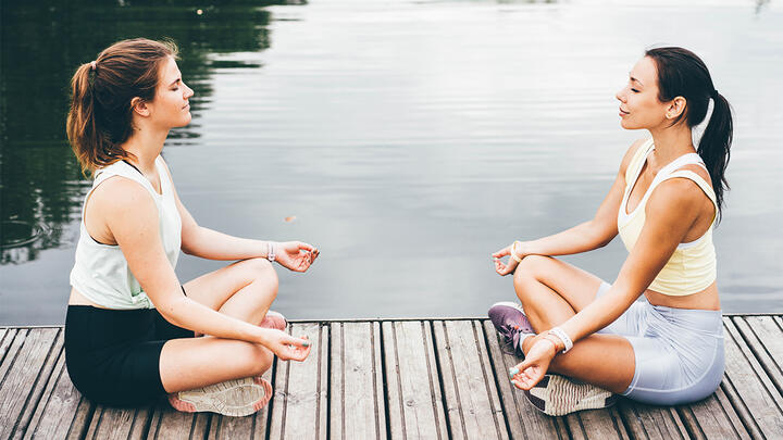 Light exercising during your period might alleviate period cramps. A photo of two young woman sitting on a boardwalk by the lake, meditating